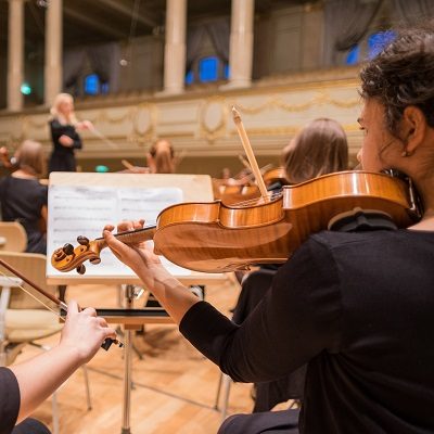 An artistic picture of a girl playing the violin, looking out onto the conductor. Symbolises the services for education concerts