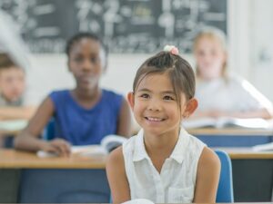 Image of a little girl smiling in class with a book in front of her. Symbolises the Services For Education SCITT course