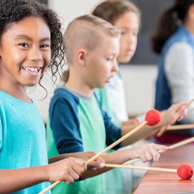 Picture of a girl playing some drums in class, smiling at the camera. Symbolising the work that the Services For Education Friends does