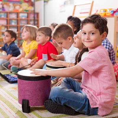 Children in a classroom playing drums, sat on the floor. Child smiling into camera. Symbolises Inspiring Sounds