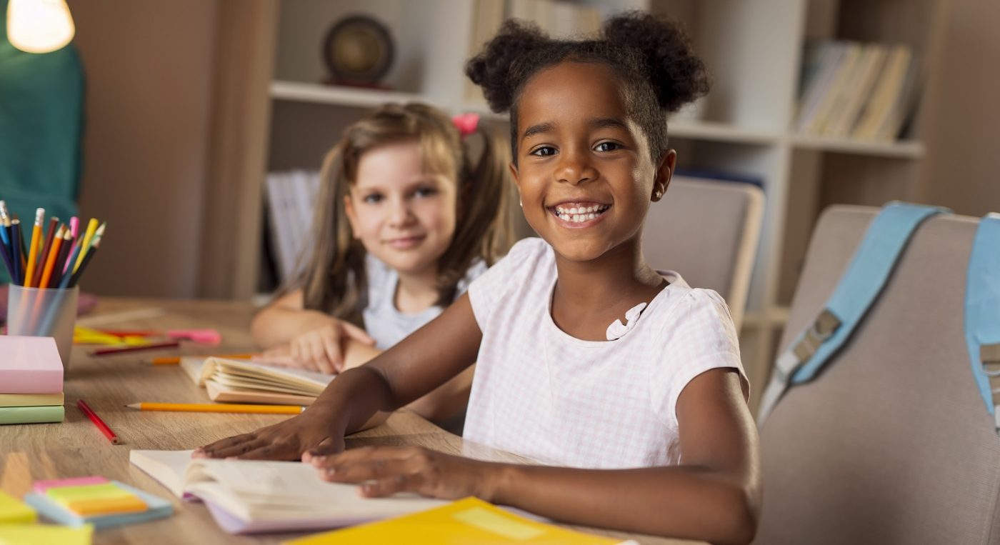 Two little girls smiling at the camera as they write in workbooks. Making Learning Stick - Curriculum 2019
